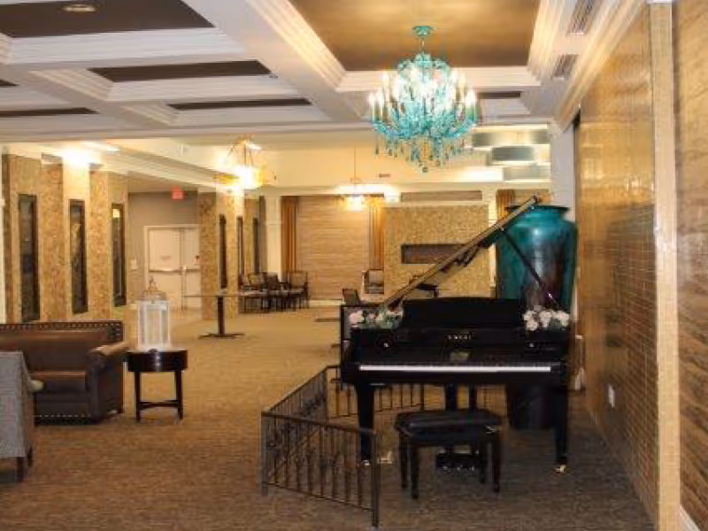 Interior view of a senior living facility common area featuring a black grand piano with a matching bench, a large decorative vase, a turquoise chandelier, several chairs and tables, and a seating area with sofas. The space has a carpeted floor and warm lighting.