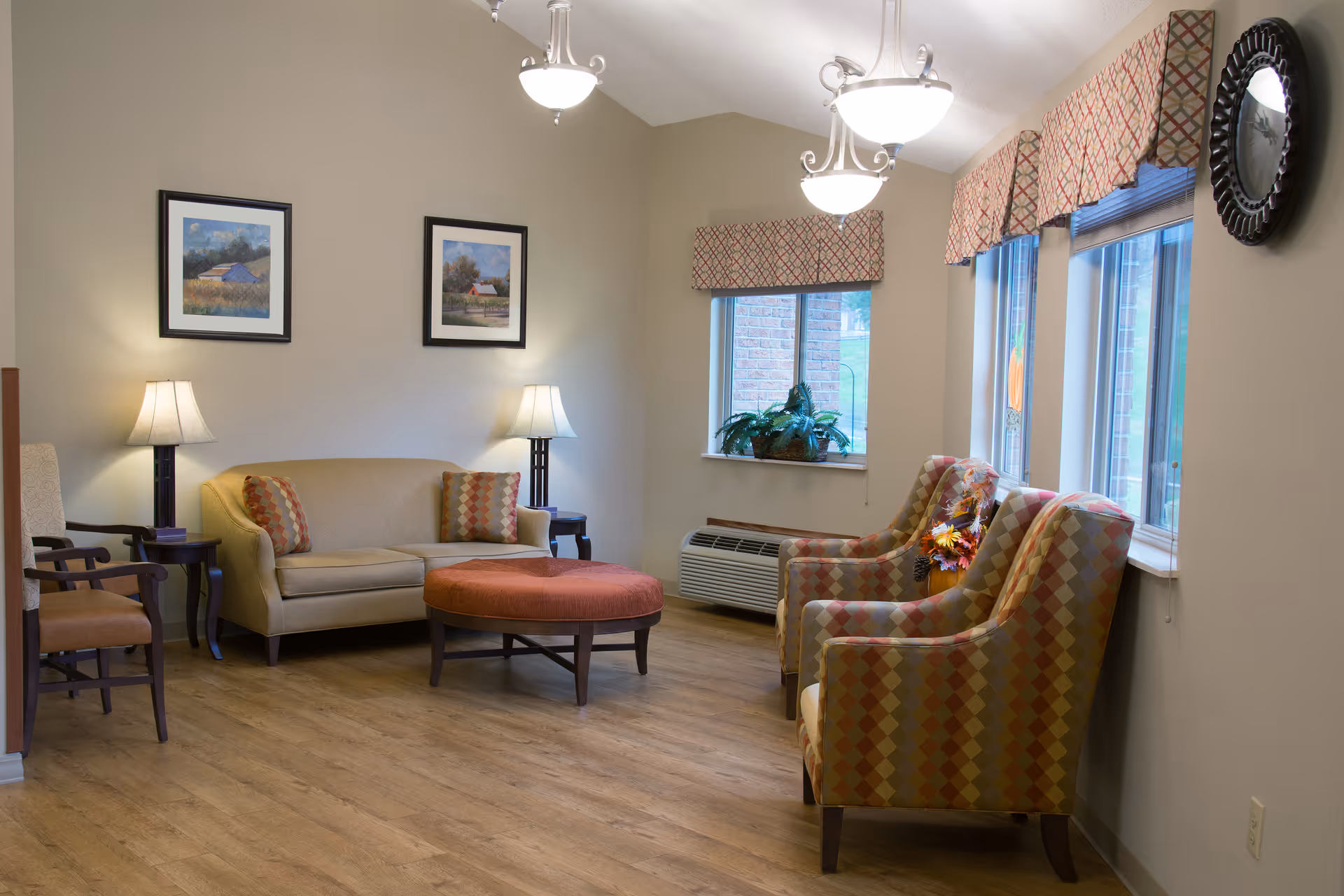 A cozy sitting area in a senior living facility with a beige sofa, two patterned armchairs, a round orange ottoman, two side tables with lamps, framed landscape paintings on the wall, and three windows with patterned valances letting in natural light.