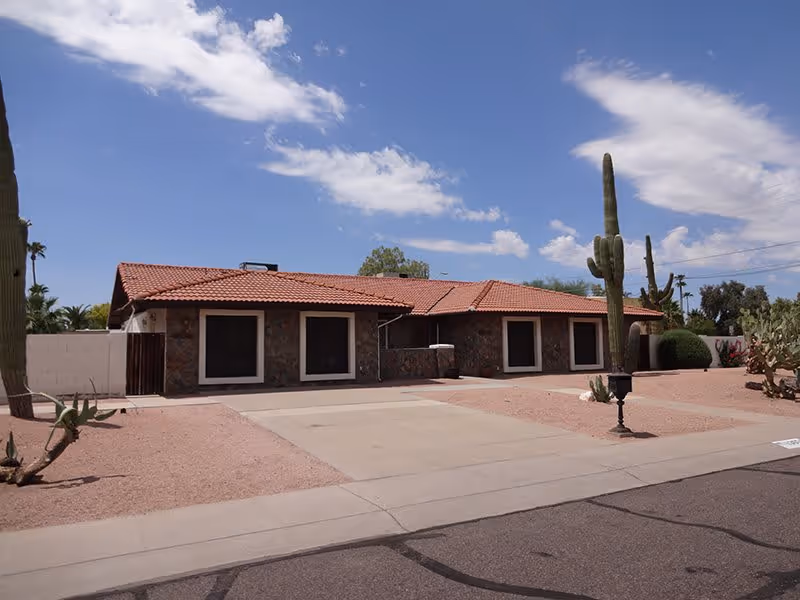Single-story residential building with a red tile roof and stone exterior walls under a partly cloudy blue sky. The front yard is landscaped with desert plants including tall cacti and gravel, and there is a concrete driveway leading to the house.
