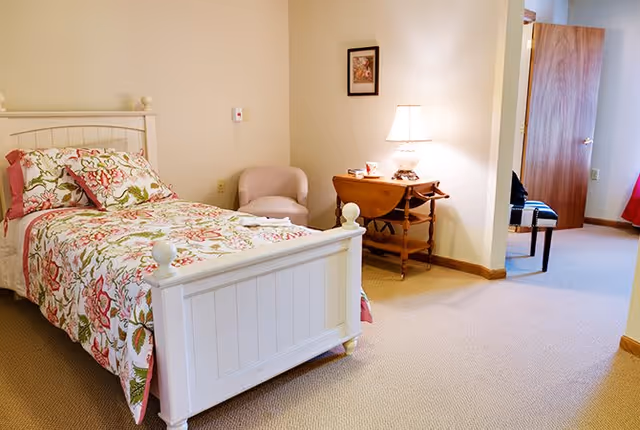 A cozy bedroom with a white wooden bed frame featuring floral bedding in shades of pink, green, and white. Next to the bed is a small beige armchair and a wooden side table with a lamp, a cup, and some items on it. The room has beige carpet and cream-colored walls. An open door leads to another room with a chair visible inside.