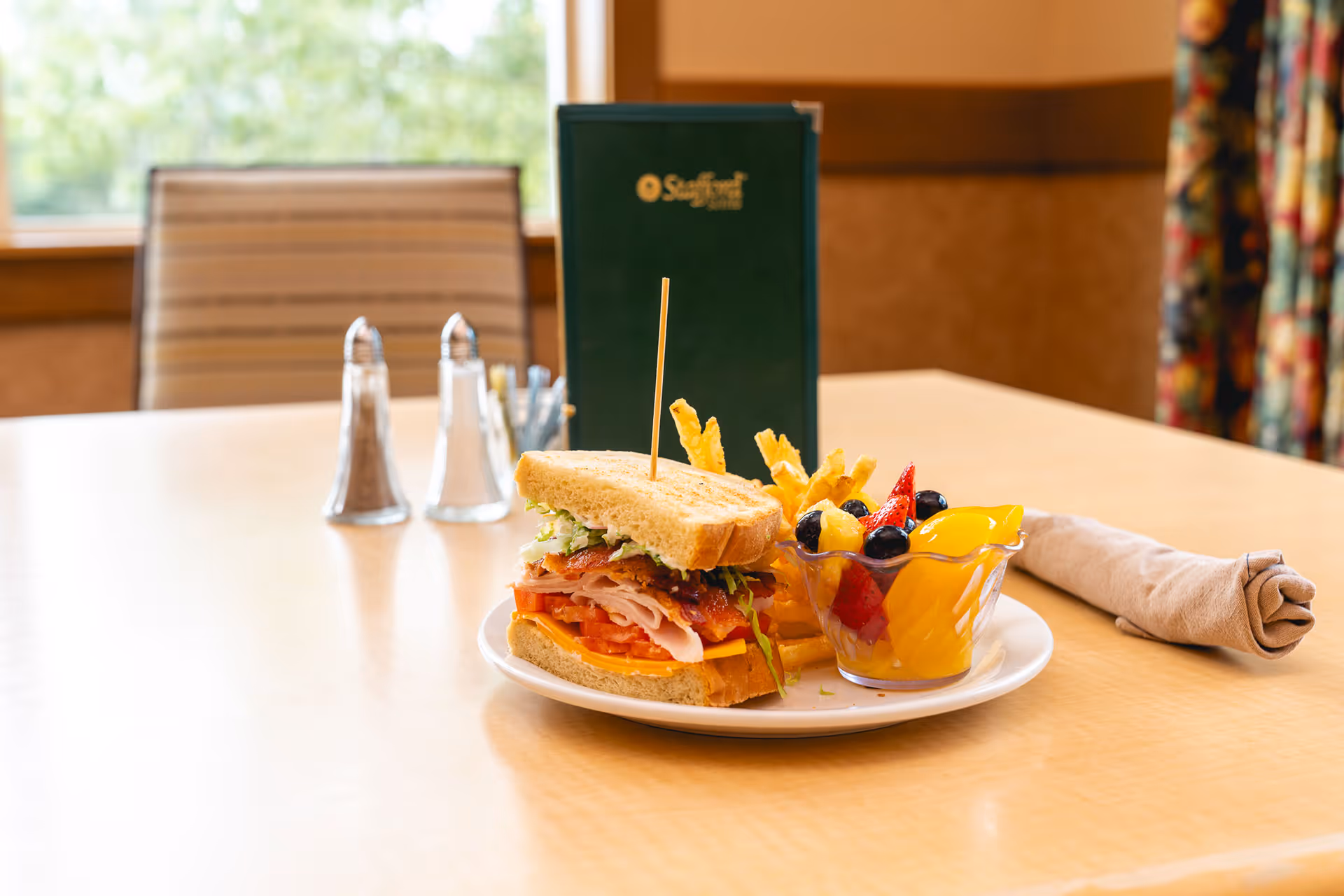 A plate with a sandwich, French fries, and a cup of mixed fruit on a wooden table in a dining area. Behind the plate, there is a green menu standing upright, salt and pepper shakers, and a rolled beige napkin. A window and a striped chair are visible in the background.