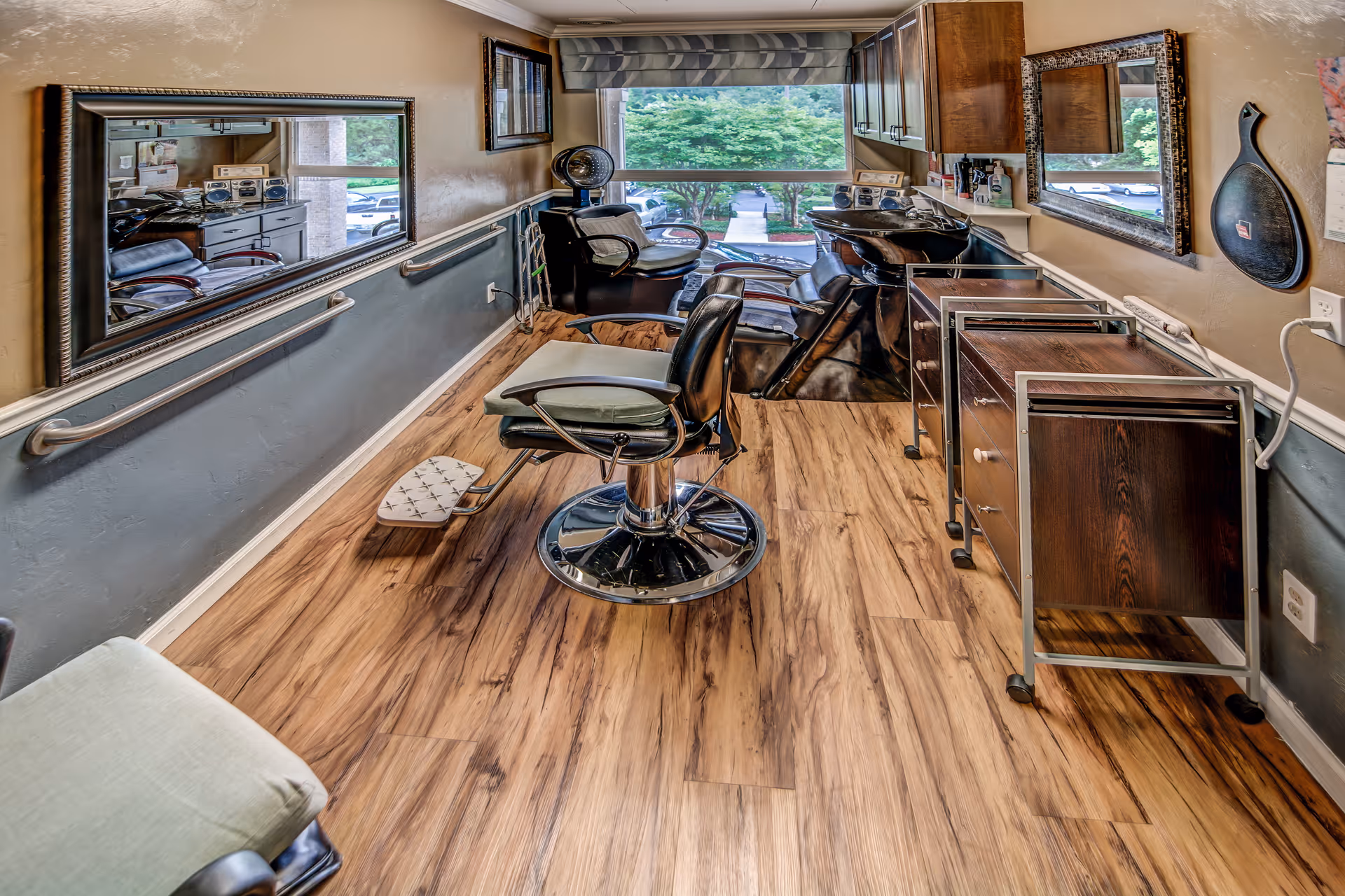 Interior view of a hair salon area with wooden flooring, a salon chair with footrest, a hair washing station with reclining chairs, large mirrors on the walls, and a window showing greenery outside.