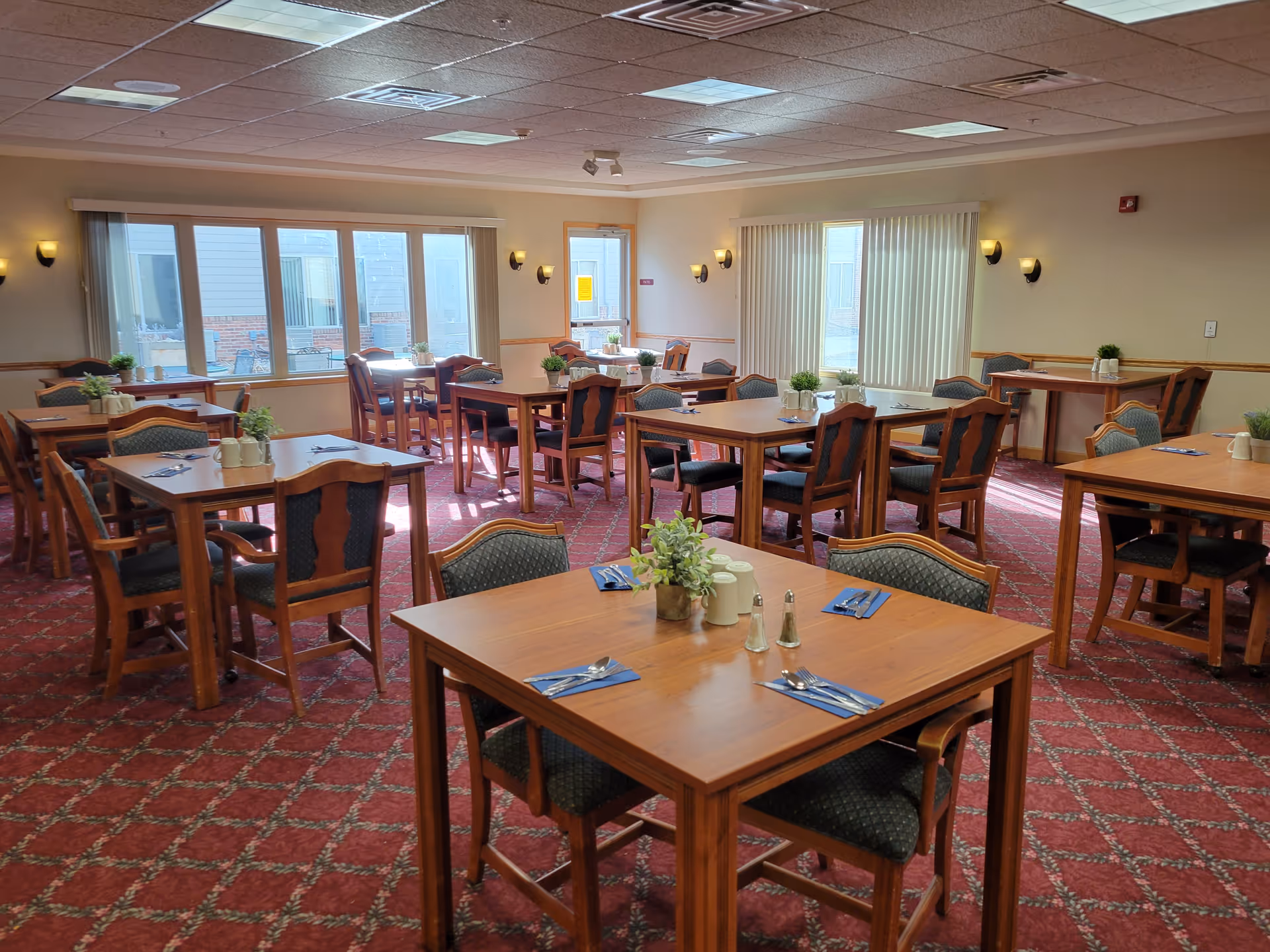 A well-lit dining room with multiple wooden tables and chairs arranged neatly. Each table has place settings with utensils on blue napkins, salt and pepper shakers, and small potted plants as centerpieces. Large windows and glass doors allow natural light to fill the room, which has a red patterned carpet and beige walls with wall-mounted lights.