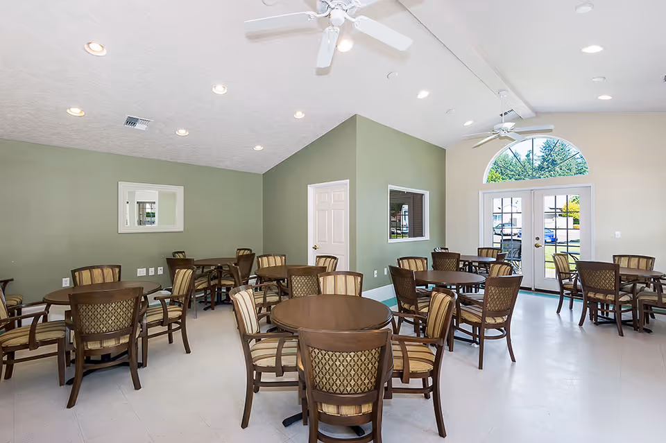 Bright dining/activity room with multiple round tables and upholstered chairs, a green accent wall, ceiling fans, and glass doors with an arched window.