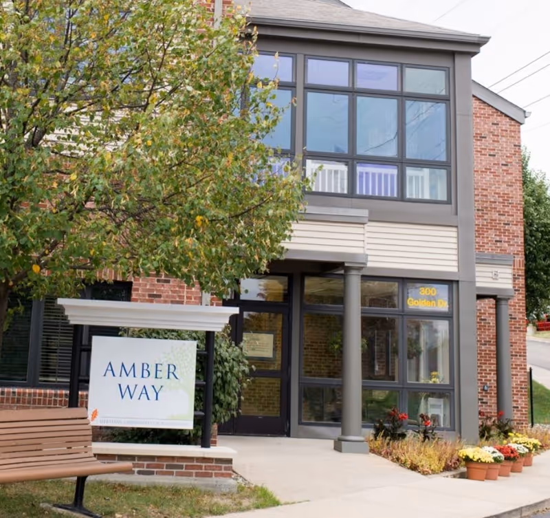 Exterior view of Amber Way facility showing a brick building with large windows, a tree with green leaves, a wooden bench, and a sign that reads 'Amber Way Heritage Community of Kalsmpton'. There are also potted plants with flowers near the entrance.
