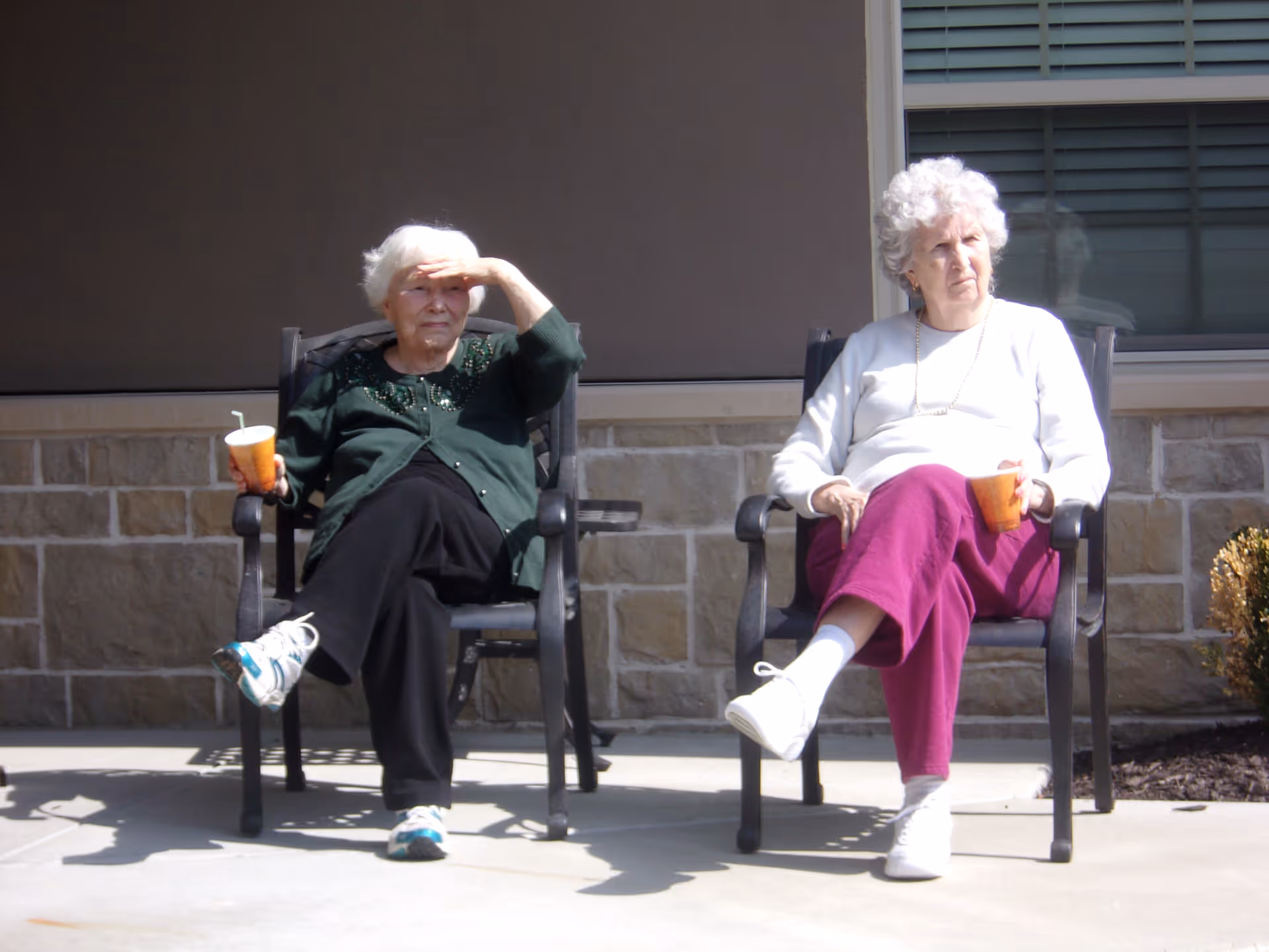 Two elderly women sitting outdoors on black chairs against a stone and beige wall. One woman is wearing a green cardigan and black pants, shielding her eyes from the sun with one hand and holding a cup with a straw in the other. The other woman is wearing a white sweater and magenta pants, holding an orange cup and looking to the side. Both are seated on a concrete patio with some small bushes visible on the right side.