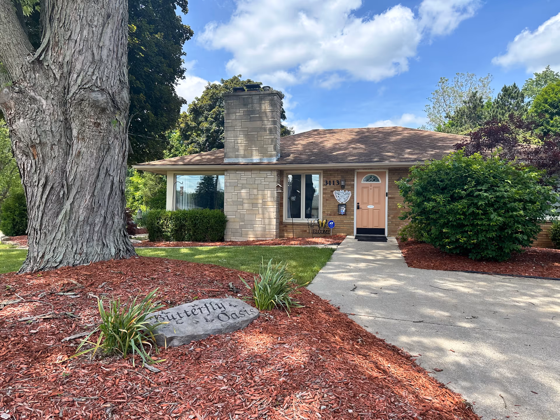 Single-story brick building with a chimney, a peach-colored front door, and a concrete walkway leading to the entrance. The building is surrounded by well-maintained landscaping with mulch, bushes, a large tree, and a stone sign that reads 'Butterfly Oasis'. The sky is partly cloudy.