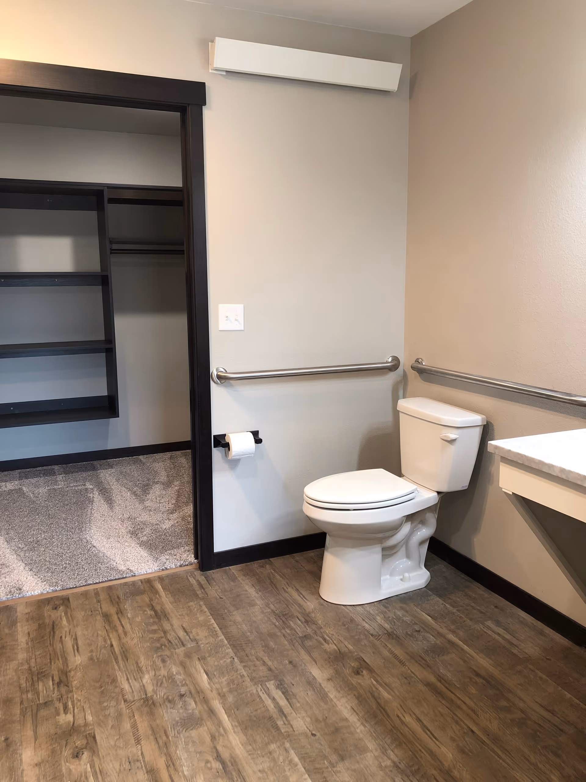 A clean, accessible bathroom with a white toilet, stainless steel grab bars on the walls, a toilet paper holder with a roll of toilet paper, and a countertop on the right side. The floor has wood-like vinyl flooring, and there is an open doorway leading to a carpeted closet with shelves and hanging rods.