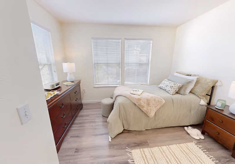 Bright bedroom with a neatly made twin bed, two windows, a dresser and a nightstand with lamps.