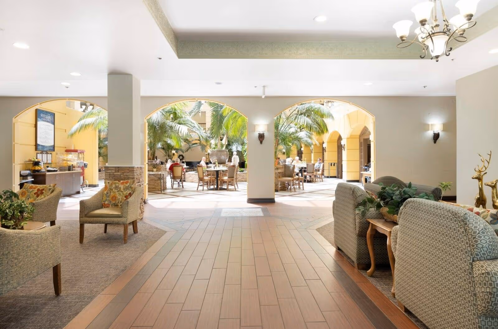Bright, spacious senior living common area with seating and dining tables opening onto a palm-lined atrium.
