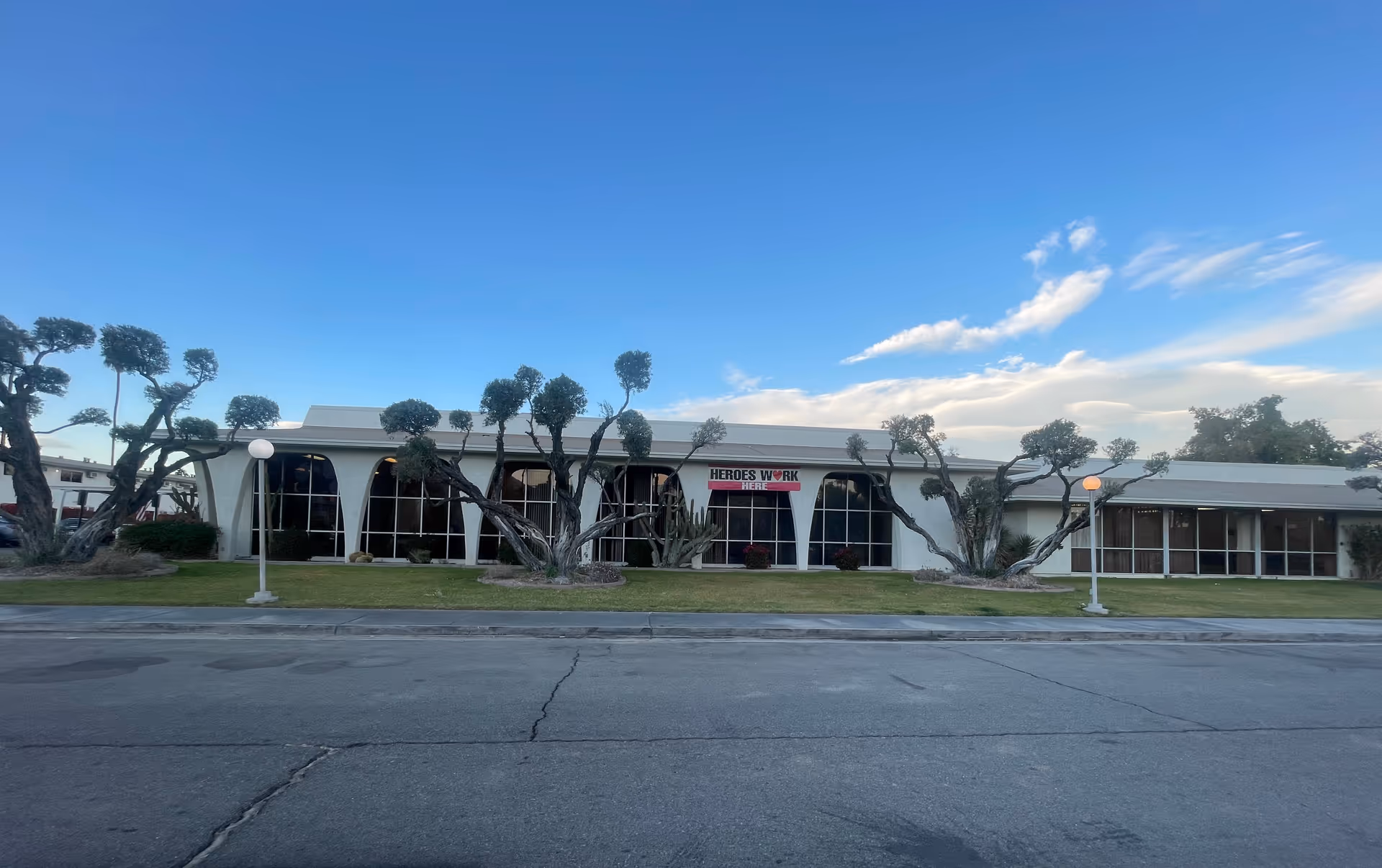 Exterior view of a single-story building with large arched windows and uniquely pruned trees in front. The building has a sign that reads 'HEROES WORK HERE'. There are two street lamps on the grassy area in front of the building, and the sky is clear with some clouds.