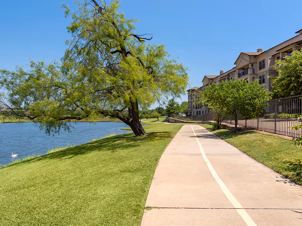 A paved walking path runs alongside a calm lake with a large tree leaning over the water on the left. On the right side, there is a grassy area with smaller trees and a multi-story senior living building in the background under a clear blue sky.