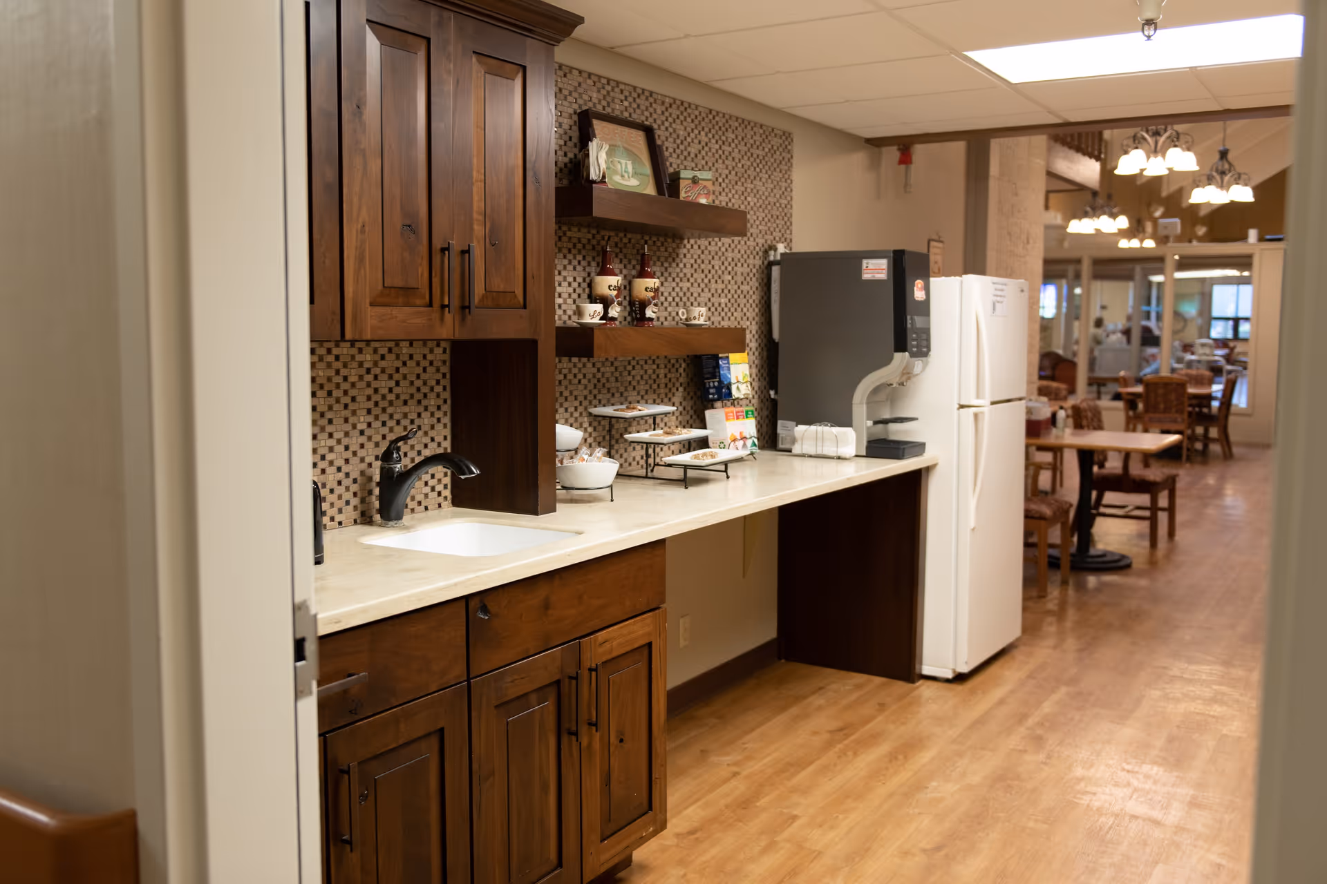 Interior view of a kitchenette area with wooden cabinets, a sink, a coffee machine, and a refrigerator. The countertop has coffee cups, bottles, and snacks. In the background, there is a dining area with tables and chairs under hanging lights.