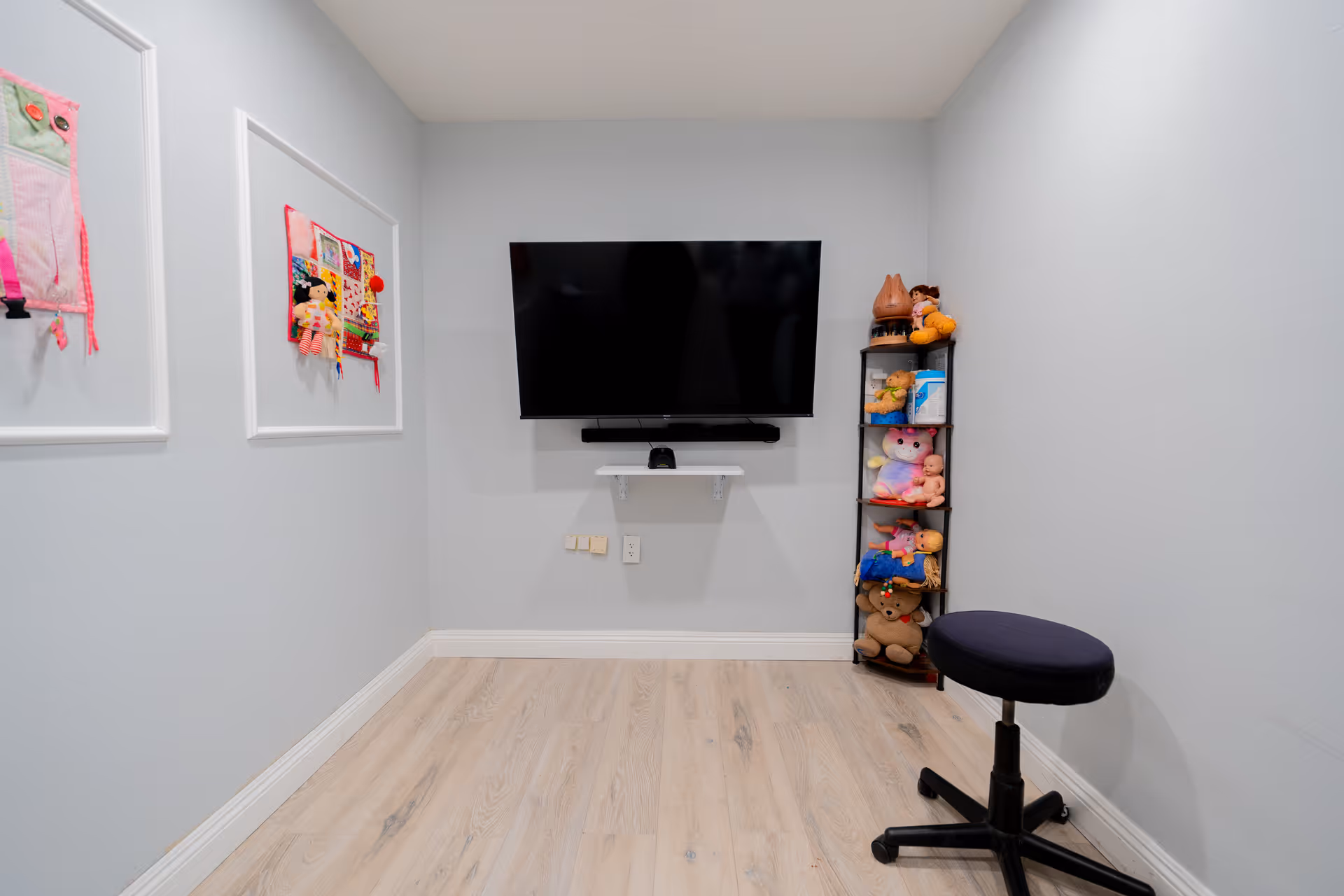 Small interior room with a wall-mounted TV, a narrow shelf of stuffed toys, framed fabric art on the wall, and a rolling stool.