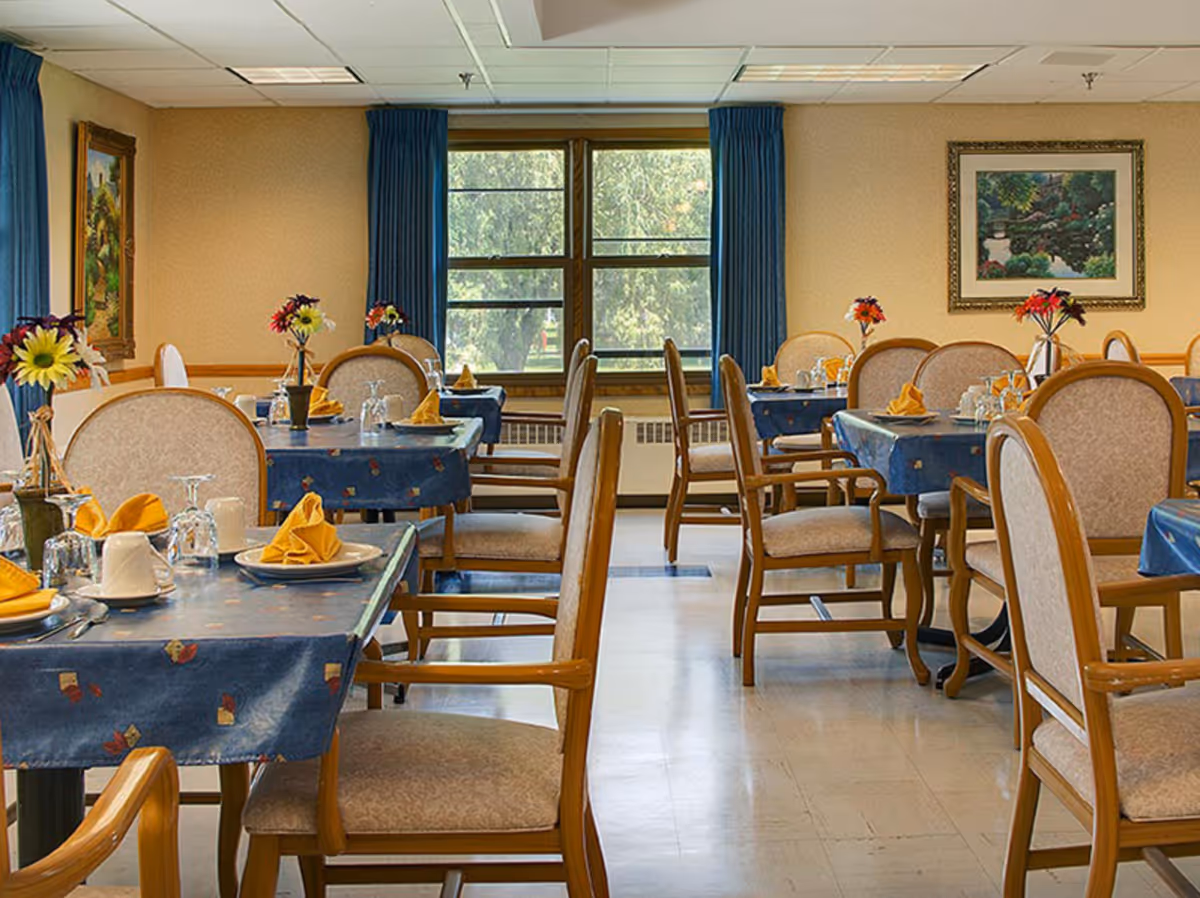 A dining room with several tables covered in blue tablecloths, each set with plates, cups, glasses, and yellow folded napkins. Chairs with wooden frames and cushioned seats surround the tables. There are flower vases on each table, and large windows with blue curtains let in natural light. Paintings hang on the beige walls.