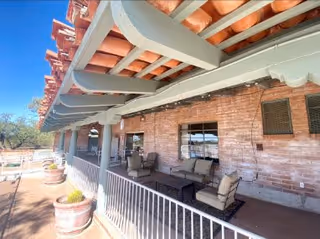 Outdoor patio area with cushioned seating and tables under a covered roof with exposed beams, adjacent to a brick building wall with windows and ventilation panels. Several large potted plants line the railing along the patio.