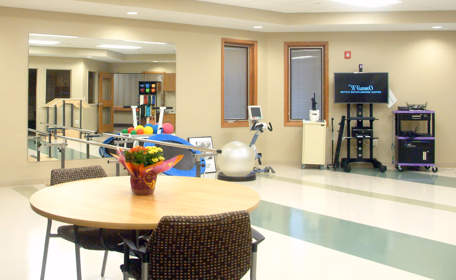 A therapy or rehabilitation room with a round wooden table and four chairs in the foreground. On the table is a small potted plant with yellow flowers. In the background, there are exercise and therapy equipment including parallel bars, colorful therapy balls, a stationary bike, and a large exercise ball. Two windows with blinds and a TV screen mounted on a stand are also visible. The room has light-colored walls and a clean, spacious floor with green and white tiles.