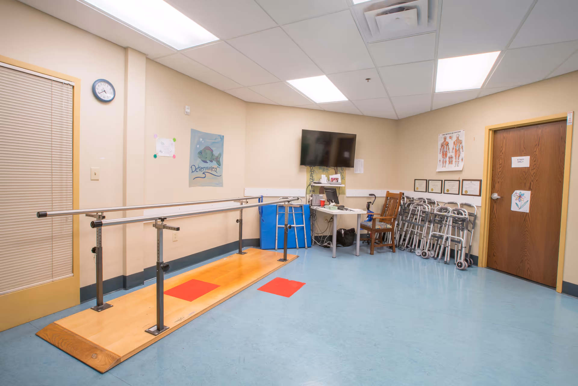 A senior living therapy room with parallel walking bars on a wooden platform, stacked walkers, a desk and TV, and medical posters on the walls.
