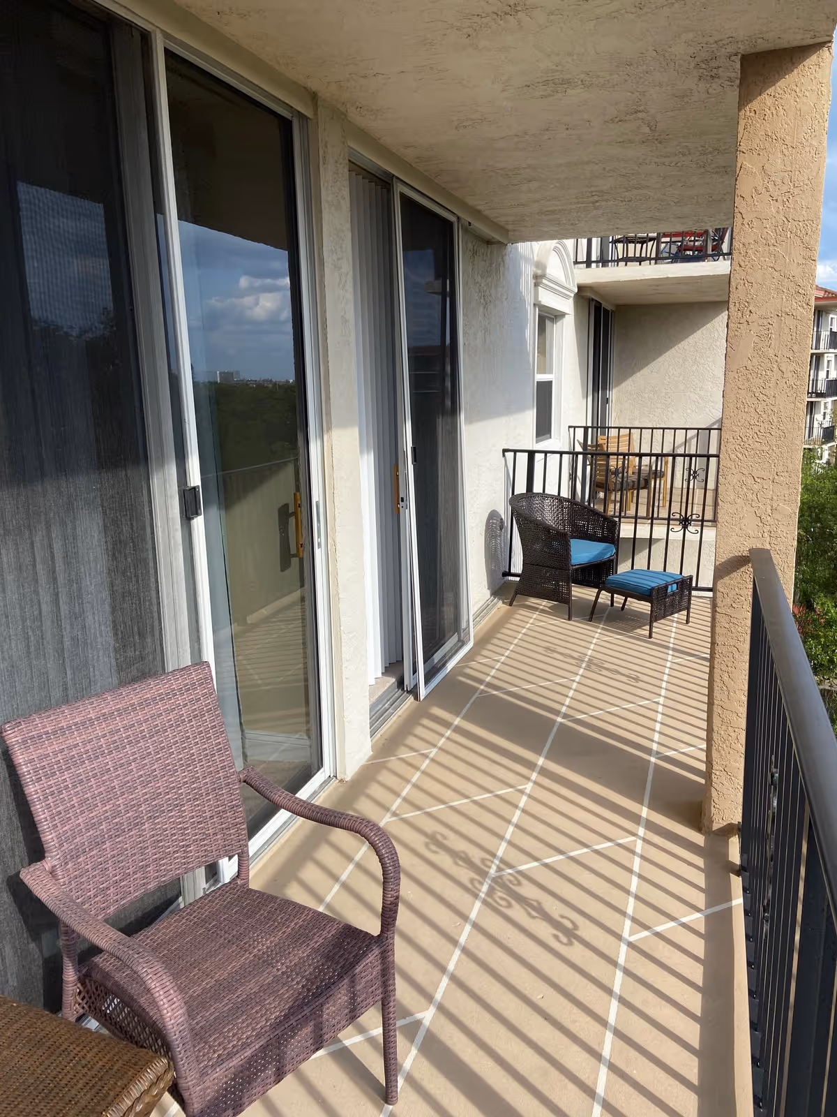 A balcony with two sets of outdoor wicker chairs and small tables. The balcony has a beige tiled floor with white grout lines and black metal railings. Sliding glass doors lead inside the building, and the balcony overlooks greenery and other buildings in the distance under a partly cloudy sky.