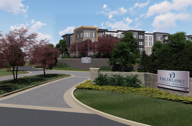 Exterior view of The Delaney at the Green senior living facility showing a modern multi-story building surrounded by landscaped greenery and trees with a curved driveway leading to the entrance. Two signs display the facility's name near the driveway.