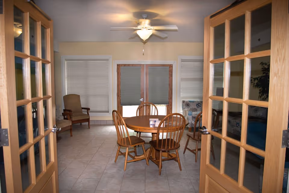 View through open wooden French doors into a room with a round wooden table and four wooden chairs. The room has tiled flooring, two windows with closed blinds, a ceiling fan with light, and additional seating including an armchair and a floral-patterned couch.