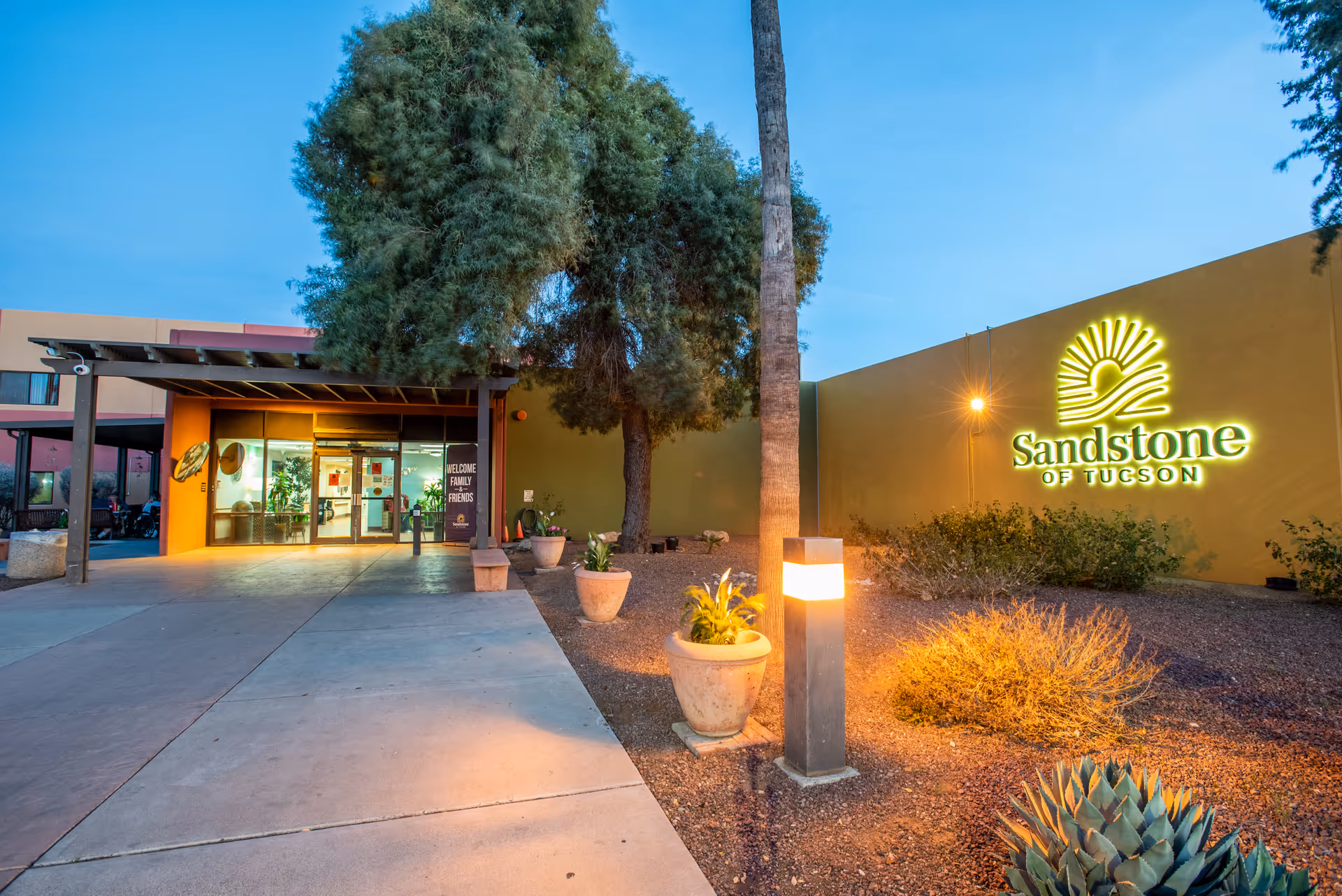 Entrance to Sandstone of Tucson senior living facility at dusk with illuminated sign, walkway, potted plants, and trees.
