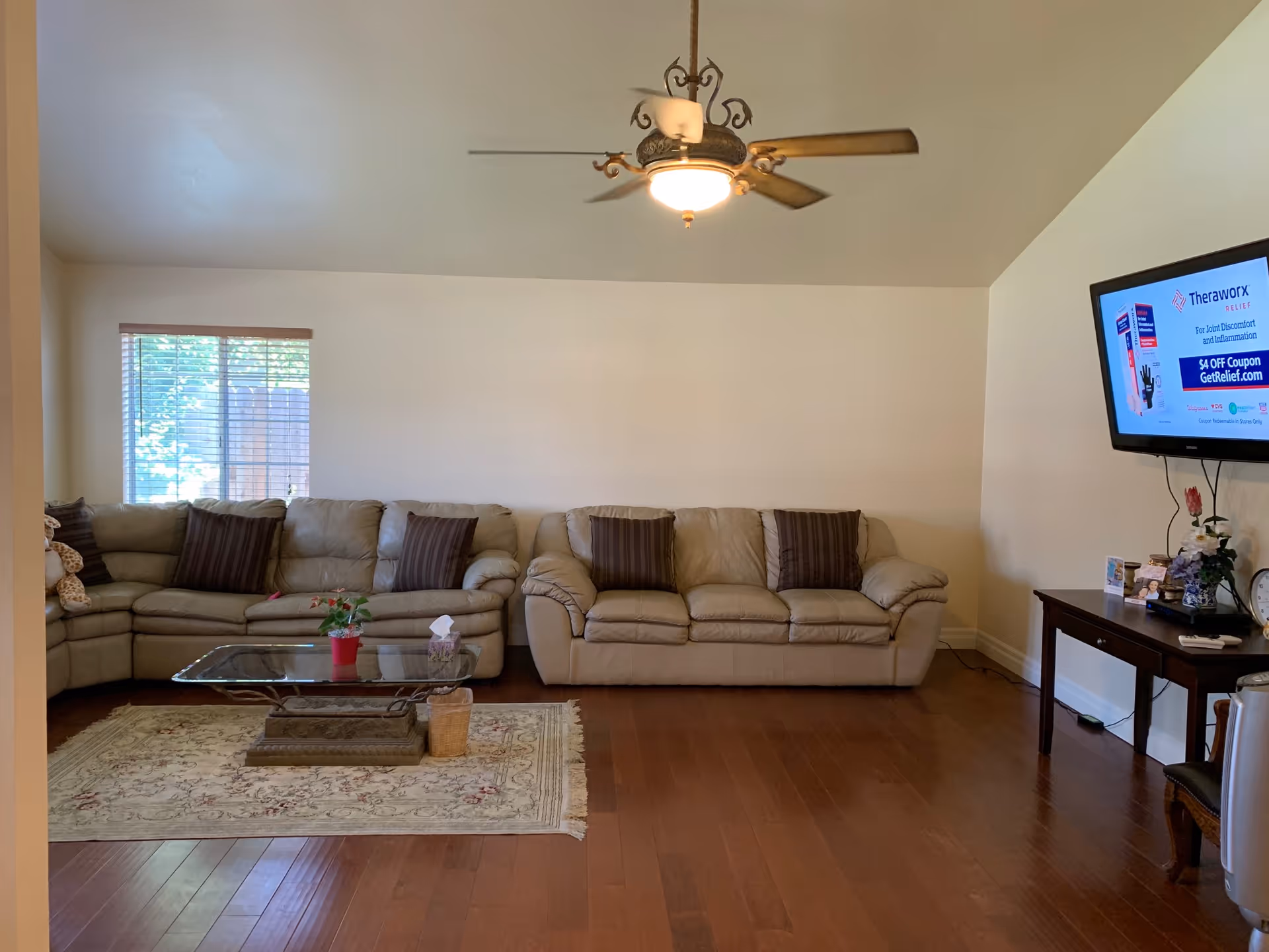 Spacious living room with beige leather sofas, a glass coffee table on a rug, a wall-mounted TV, and a ceiling fan.