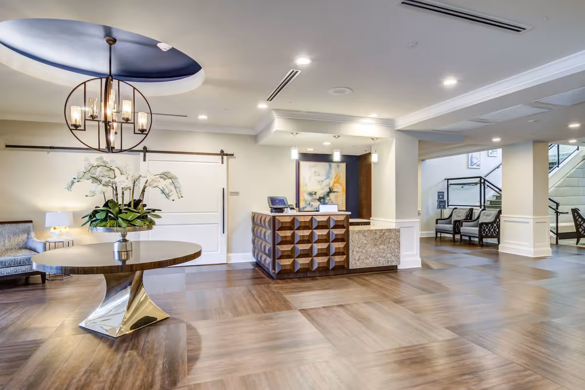 A spacious and well-lit reception area with a modern wooden reception desk, a round table with a metallic base and a floral arrangement, a chandelier hanging from a recessed ceiling, and seating areas with chairs near a staircase.