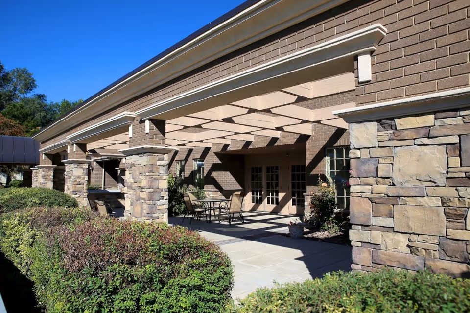 Outdoor patio area at Copper Ridge Health Care with stone pillars supporting a pergola structure, surrounded by green bushes and outdoor seating with tables and chairs under the shaded area.