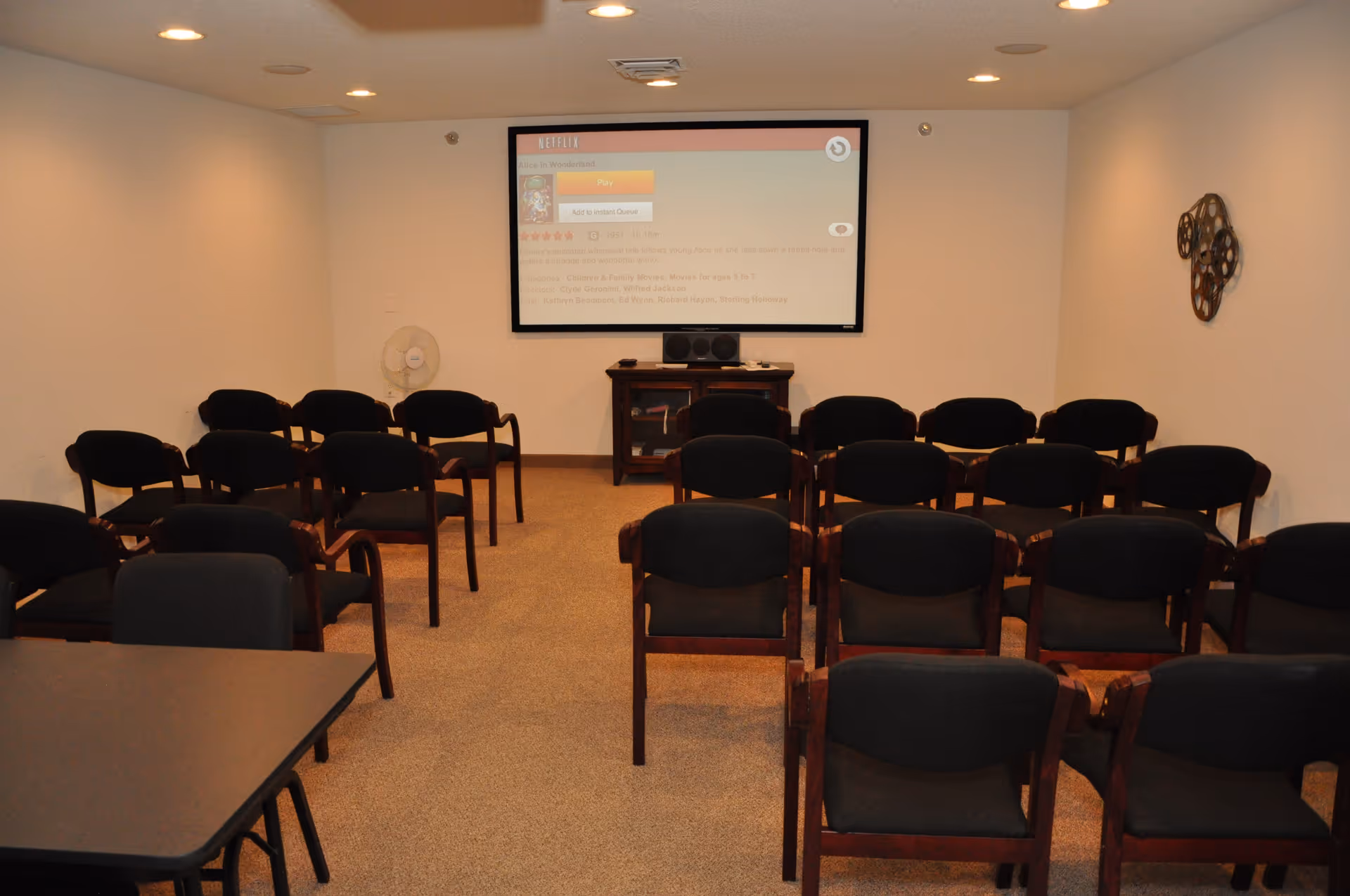 A small theater-style room with multiple rows of black cushioned chairs facing a large screen on the wall displaying a Netflix interface. The room has beige carpet, white walls, recessed ceiling lights, a small table with speakers below the screen, a standing fan in the corner, and a decorative film reel wall hanging on the right wall.