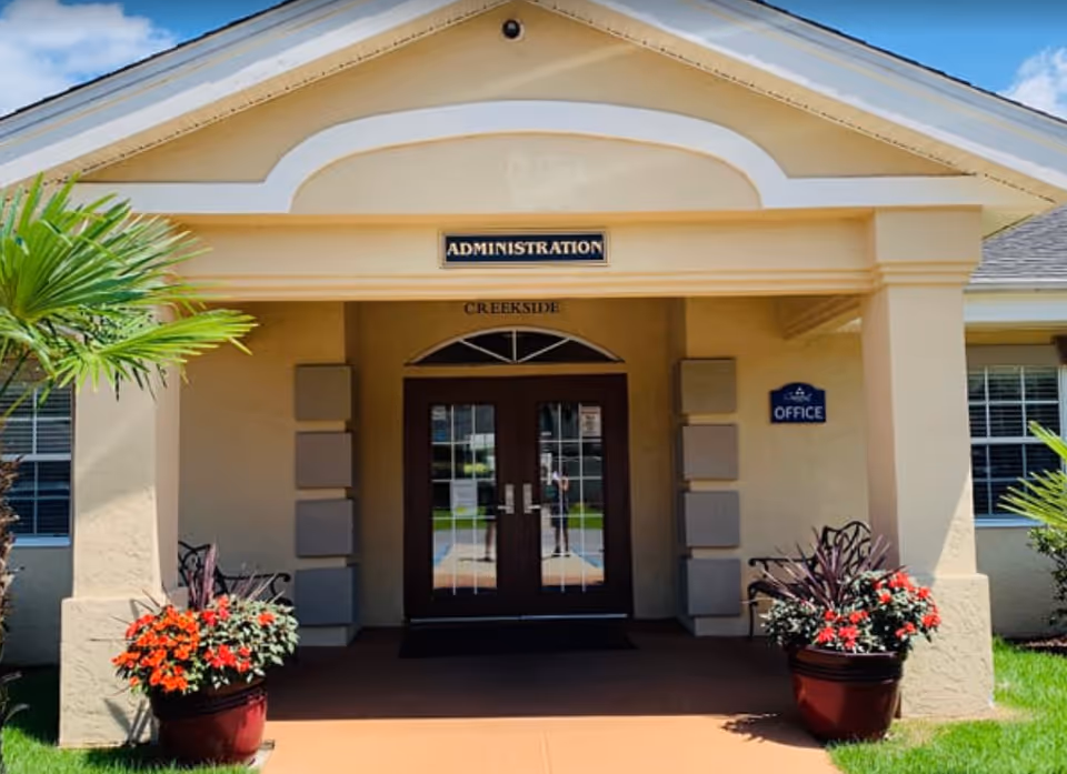 Entrance to the administration office of The Waterford at Creekside facility, featuring double glass doors under a beige portico with a sign reading 'ADMINISTRATION' above the doors. There are potted plants with red flowers on either side of the entrance and a small blue sign on the right pillar that says 'OFFICE'.