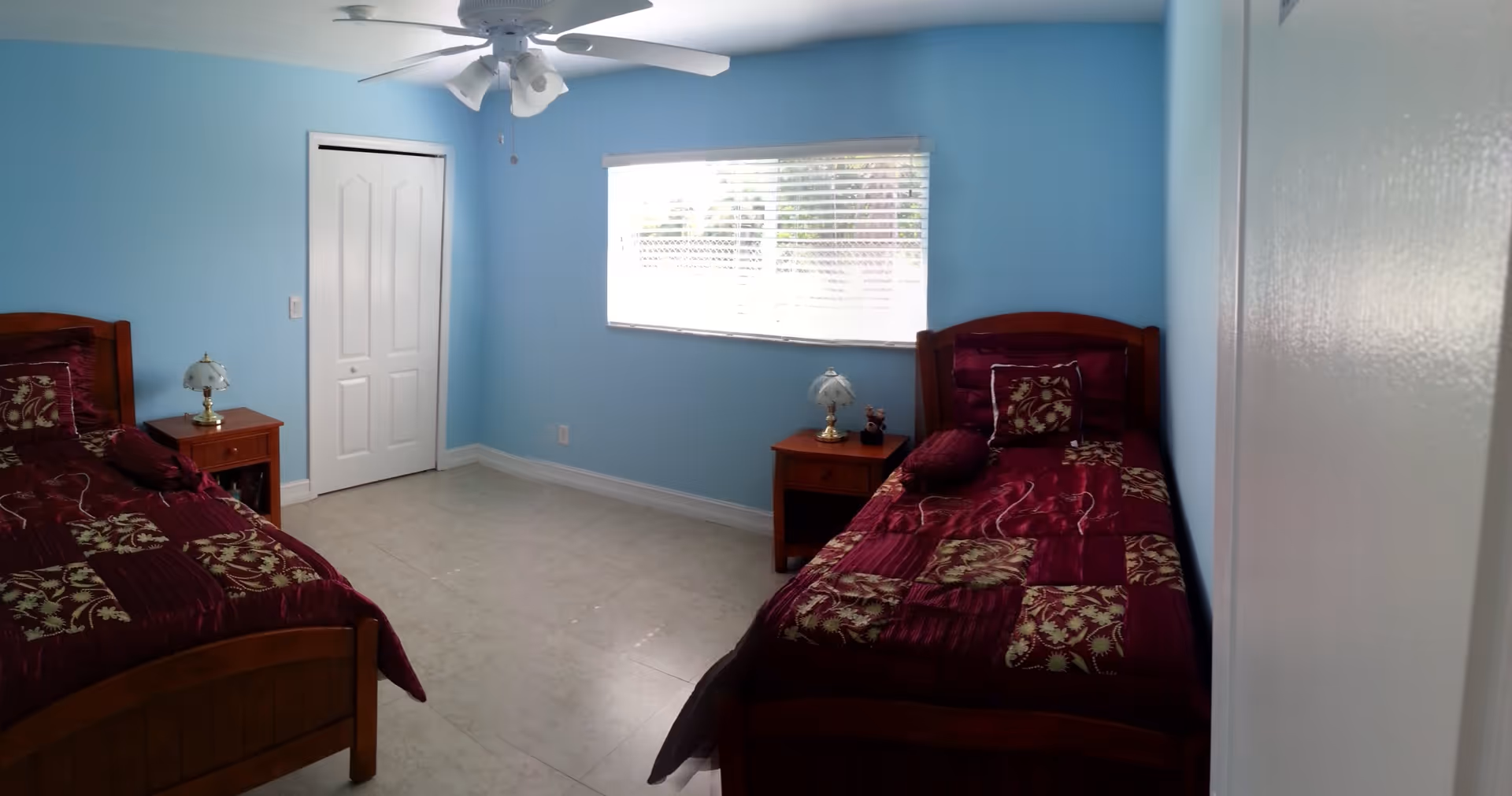 A bedroom with two single wooden beds, each with a burgundy and gold patterned bedspread and matching pillows. There are two wooden nightstands, each with a small lamp. The walls are painted light blue, and there is a window with white blinds letting in natural light. A white ceiling fan with lights is mounted on the ceiling, and a white closet door is visible on the left wall.