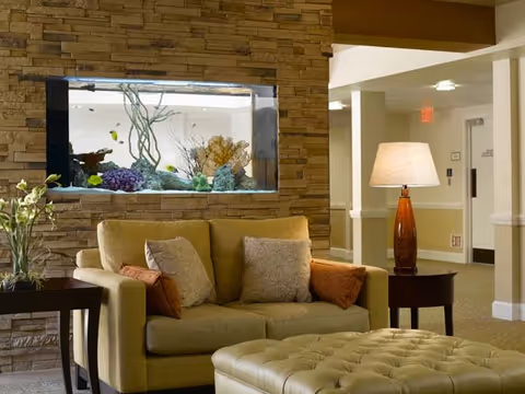 Comfortable seating area with a loveseat, tufted ottoman, side tables, lamp and a built-in aquarium in a stone wall.