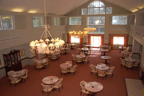 Spacious dining room with multiple round tables set for meals, chandeliers, and large windows under a high ceiling.