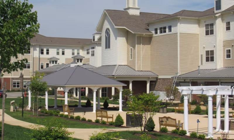 Outdoor view of Lutheran Senior Life Passavant Community featuring a large multi-story building with beige siding and white trim. In the foreground, there is a covered pavilion with a gray roof supported by white columns, surrounded by landscaped gardens, walking paths, benches, and small trees and shrubs.