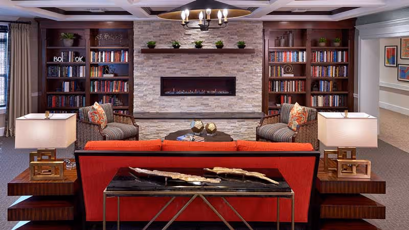 A cozy living room area featuring a red sofa facing a modern stone fireplace with a built-in electric fire. On either side of the fireplace are wooden bookshelves filled with books and decorative plants. Two striped armchairs with floral cushions flank a round wooden coffee table in front of the fireplace. Two table lamps with white shades sit on wooden side tables beside the sofa. The room has a warm and inviting atmosphere with neutral walls and carpeted flooring.