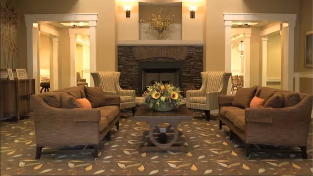 A cozy living room with two brown sofas facing each other, two striped armchairs, a wooden coffee table with a floral centerpiece, and a stone fireplace in the background. The room has warm lighting and patterned carpet with leaf designs.