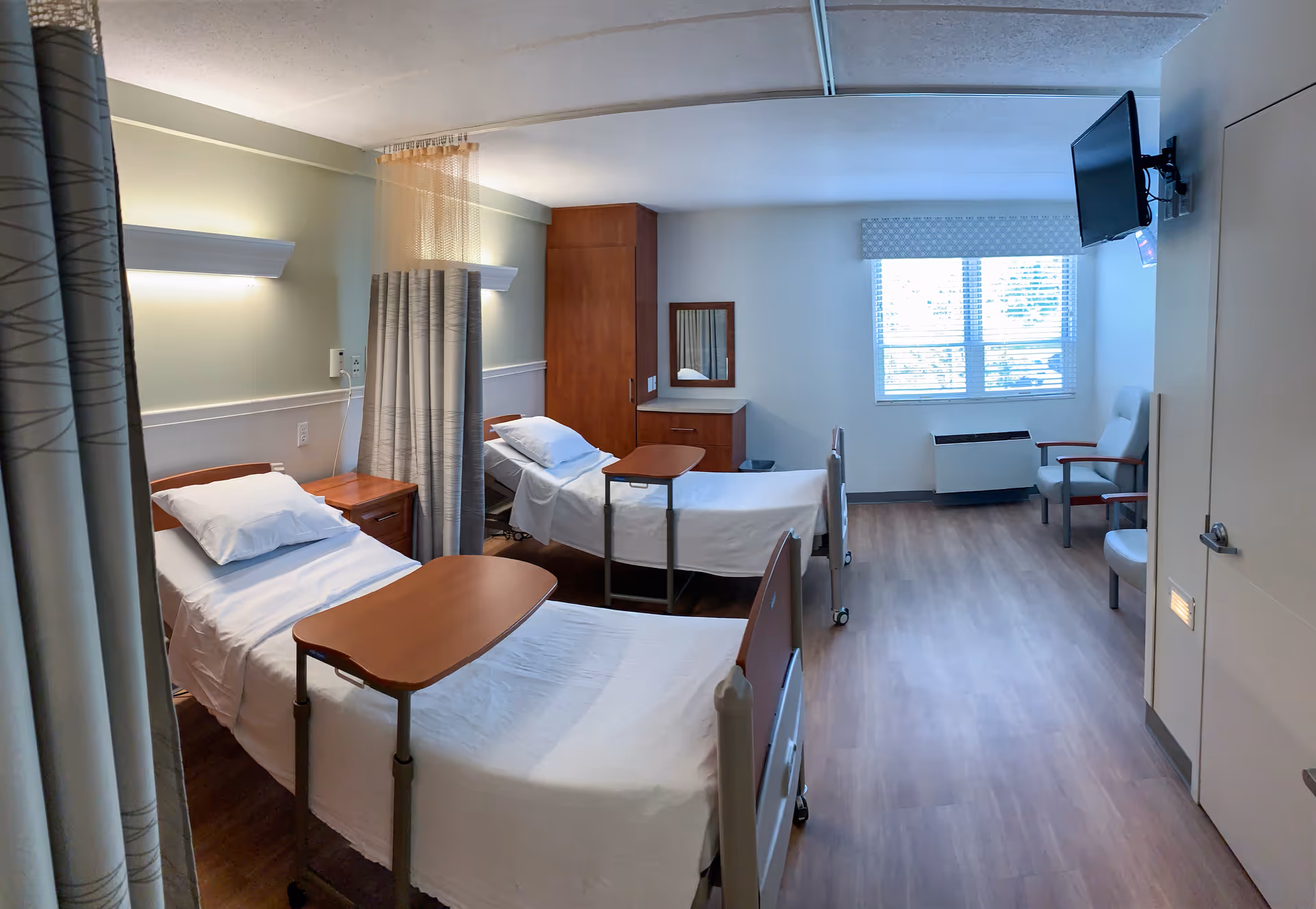A clean and well-lit room in a skilled nursing facility with two hospital beds, each equipped with a wooden overbed table. The room has light-colored walls, wood flooring, a window with blinds, two armchairs, a wall-mounted TV, and a wooden cabinet with a mirror.