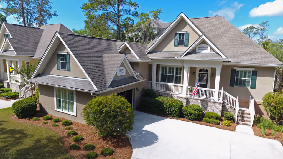 Exterior view of a beige single-family home with a gabled roof, green shutters, a front porch with white railings, an American flag, and well-maintained landscaping including bushes and a small tree. The driveway is concrete and the sky is clear with a few clouds.
