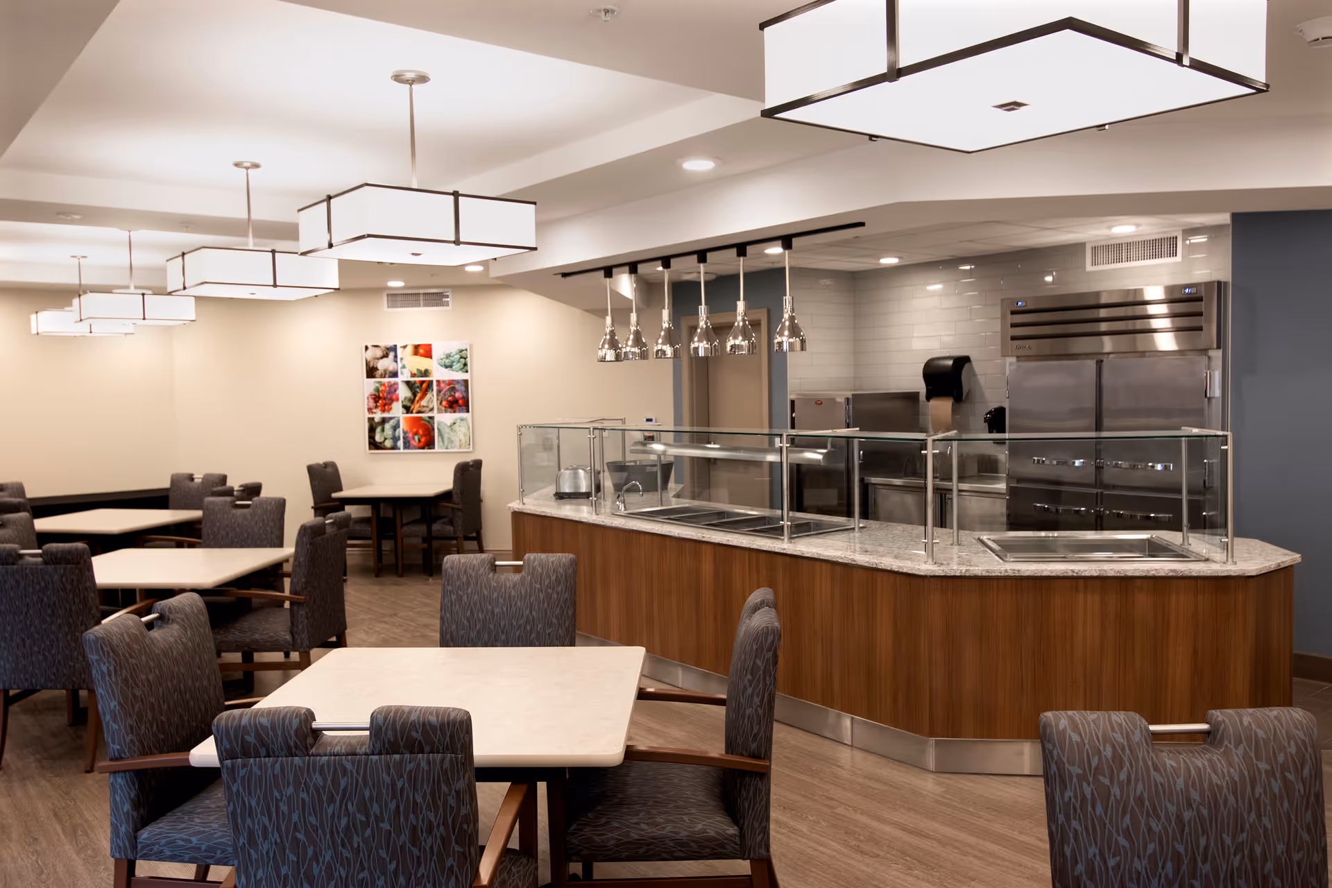A modern dining area in a senior living facility with multiple tables and cushioned chairs arranged neatly. The room features a serving counter with a glass sneeze guard and stainless steel kitchen appliances in the background. Contemporary pendant lights hang from the ceiling, and a colorful food-themed artwork is displayed on the far wall.