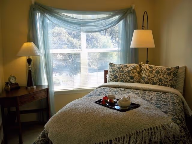 A cozy bedroom with a bed covered in patterned bedding and a fluffy white throw blanket. There is a tray with a teapot and cups on the bed. A large window with sheer blue curtains lets in natural light. On the left side of the bed is a wooden nightstand with a lamp and an alarm clock, and on the right side is a tall floor lamp.