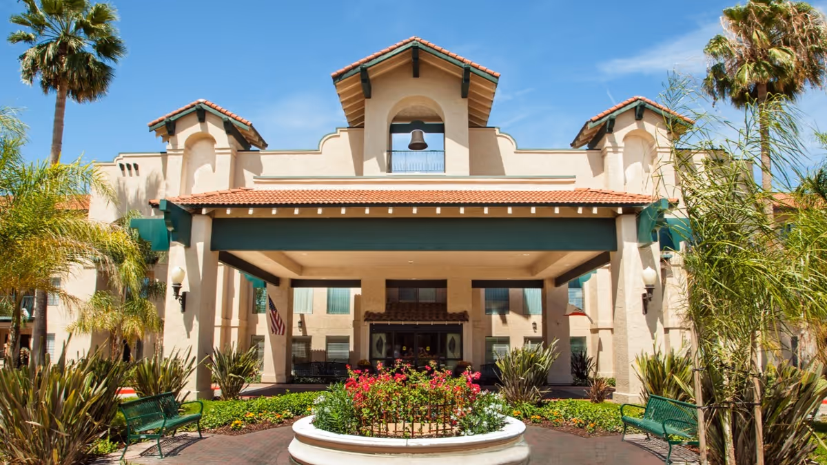 Entrance and front facade of a Mediterranean-style senior living building with a covered porte-cochere, landscaping, benches, and palm trees.