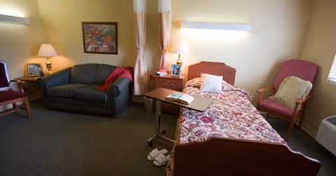 A furnished assisted-living bedroom with a single bed, floral bedspread, sofa, chairs, bedside tables and lamps.