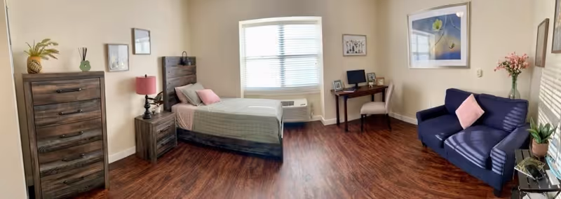 A senior living facility bedroom with a single bed featuring gray and pink bedding, a wooden nightstand with a lamp, a tall wooden dresser with decorative items on top, a small desk with a computer and chair, a blue loveseat with a pink pillow, and framed artwork on the walls. The room has wooden flooring and a window with blinds letting in natural light.