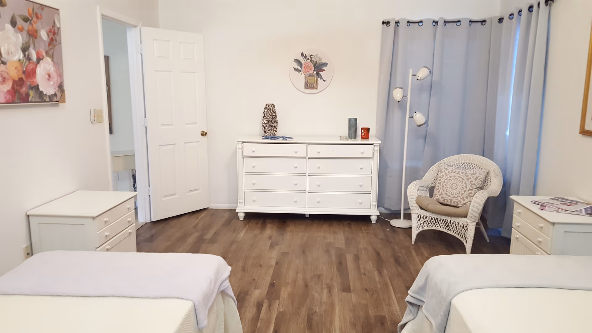A bright bedroom with two beds partially visible on either side, each with white bedding and light-colored blankets. The room features wooden flooring, white furniture including two nightstands and a dresser, a wicker chair with a cushion, a floor lamp with three lights, and light blue curtains covering a window. A floral painting hangs on the left wall and a round floral decoration is above the dresser.