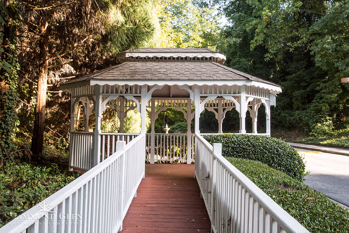A white wooden gazebo with a shingled roof situated at the end of a wooden walkway with white railings, surrounded by green bushes and tall trees in a peaceful outdoor setting.