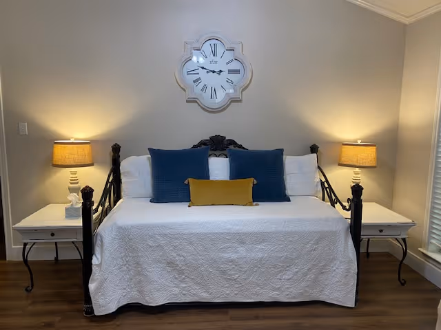 Daybed with a white cover, blue and yellow pillows, flanked by two bedside tables with lamps beneath a decorative wall clock.