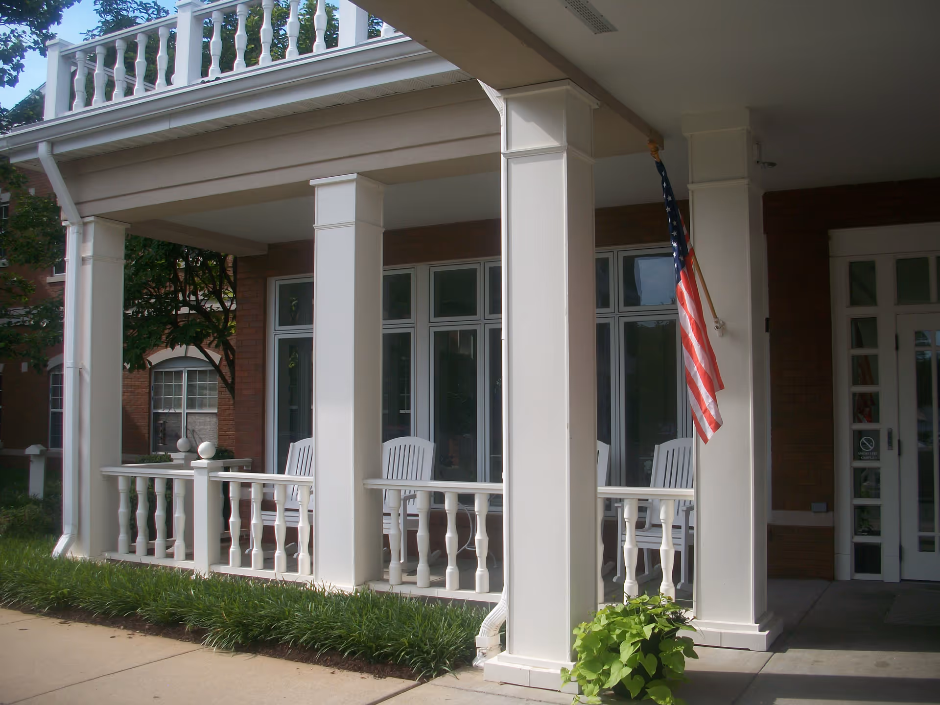 Front porch area of a building with white columns and railings, white rocking chairs, an American flag hanging from one of the columns, and greenery along the sidewalk.