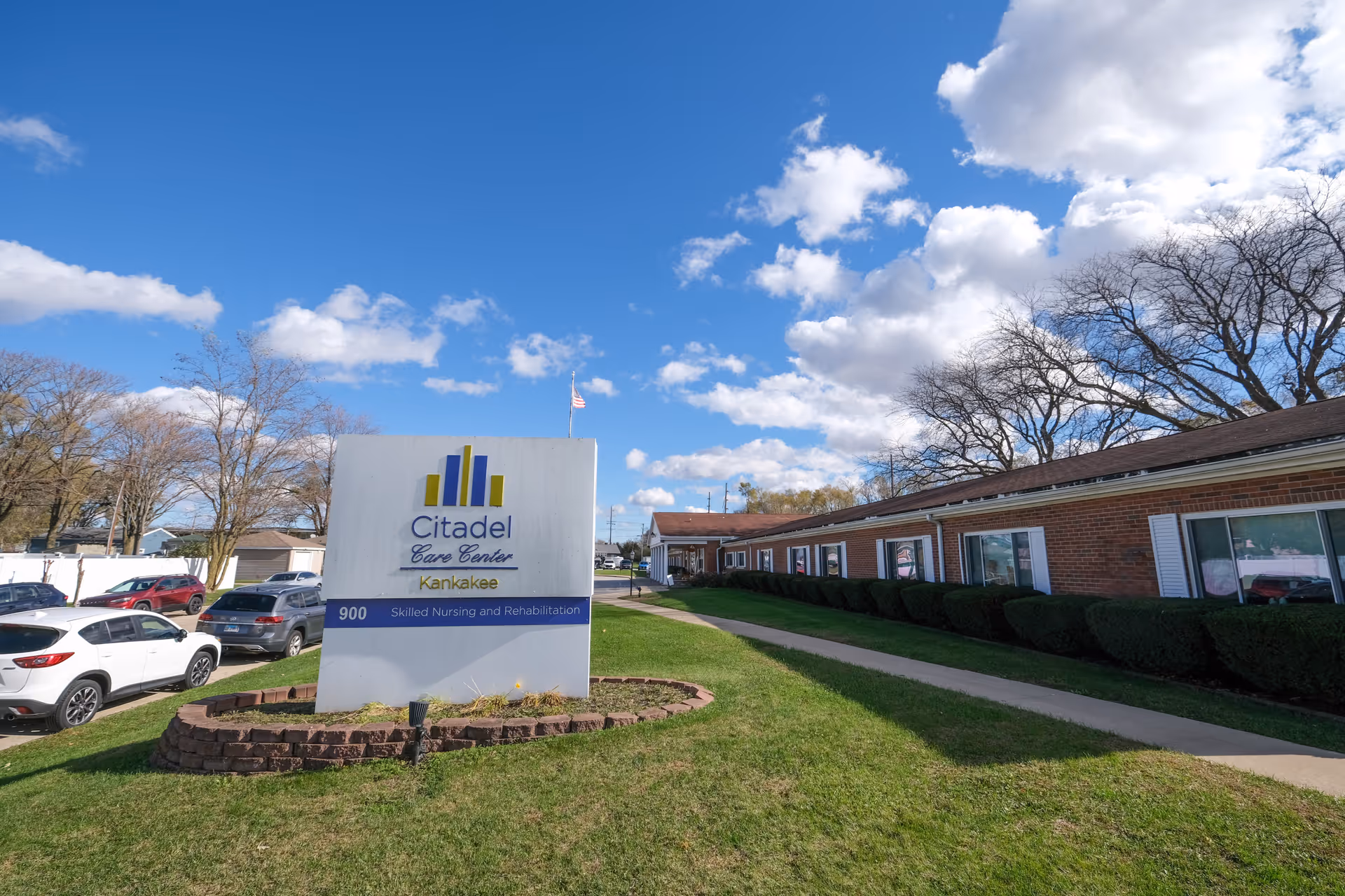 Exterior view of Citadel Care Center Kankakee, a single-story brick building with white window shutters and neatly trimmed bushes along the sidewalk. A large sign in front reads 'Citadel Care Center Kankakee, 900 Skilled Nursing and Rehabilitation.' Several parked cars are visible to the left, and the sky is blue with scattered clouds.