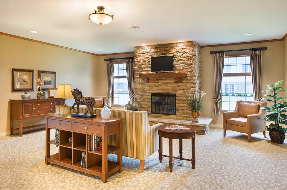 A cozy living room with a stone fireplace and a mounted TV above it. The room features two windows with curtains, a striped armchair, a leather armchair, a wooden side table, and a wooden console table with decorative items including a horse sculpture and vases. The walls are painted beige and the carpet is light-colored with a subtle pattern.